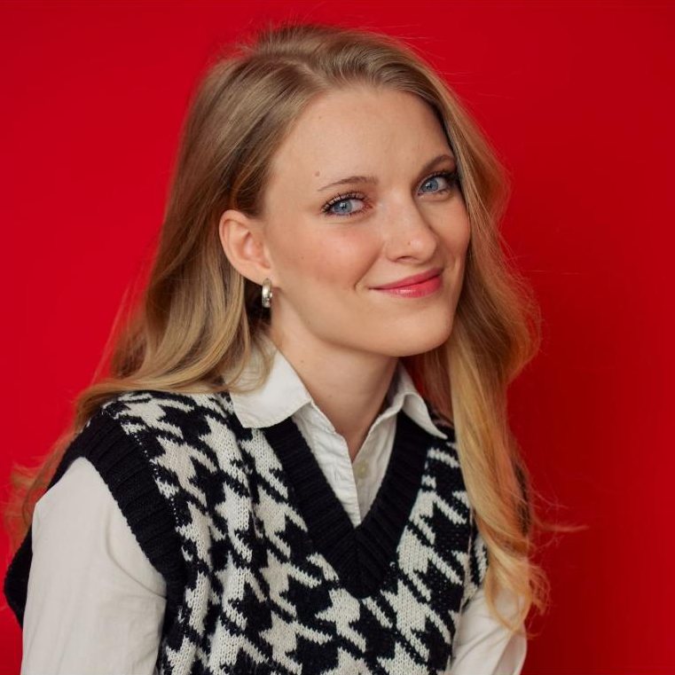Paige McKenna headshot, slight smile, houndstooth sweater vest on a red backdrop