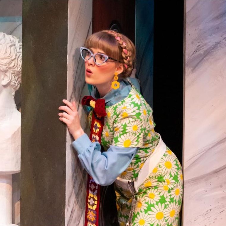 Paige as Maria in cat-eye glasses and a daisy-print dress, peeking around a column